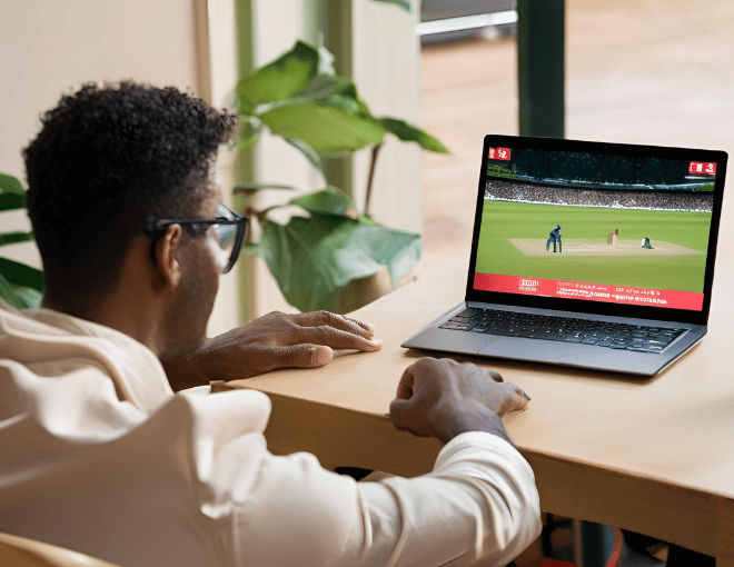 A behind-the-back shot of a man watching a cricket game on his laptop.